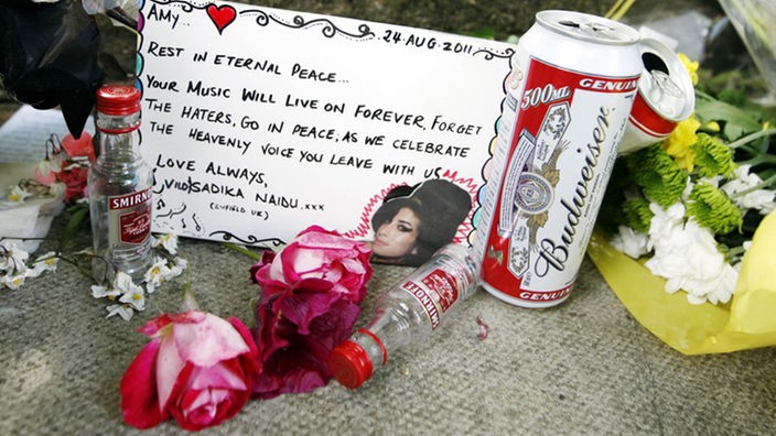Alkoholflaschen und Rosen liegen neben einer Gedenkkarte an Amy Winehouse.