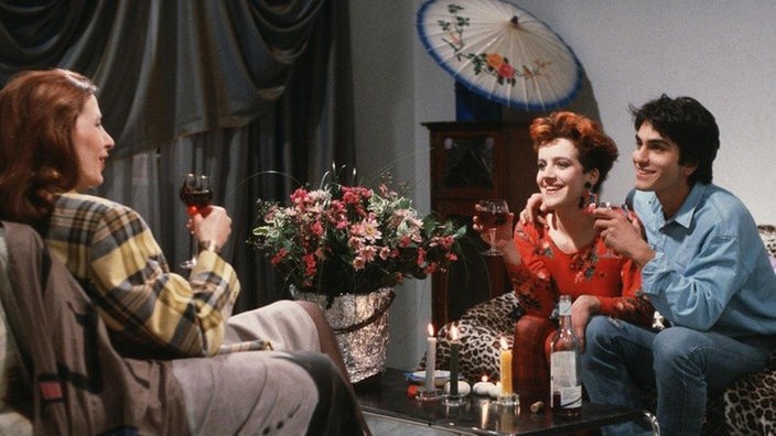 Bianca (Annette Kreft, links) stösst mit Beate (Susanne Gannott) und Vasily (Hermes Hodolides) auf deren gemeinsame Wohnung an und erzählt, dass sie die Meisterprüfung machen will.