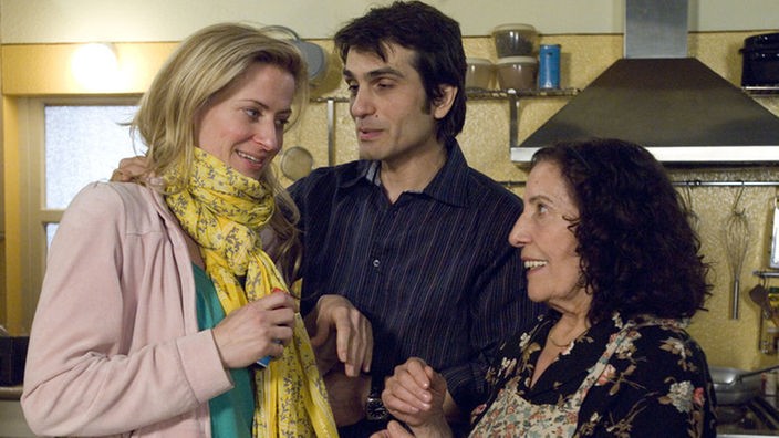 Elena (Domna Adamopoulou, rechts) und Vasily (Hermes Hodolides) bestärken Sandra (Jennifer Steffens) darin, ein Mittel gegen ihren starken Husten zu nehmen.