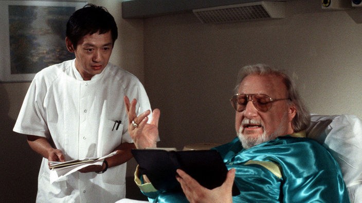 Im Krankenhaus bietet der Patient und Filmproduzent Haggerl (Walter Tschernich, rechts) dem Vietnamesen Gung (Amorn Surangkanjanajai) eine Rolle als Thailänder an.