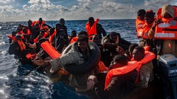 Mehrere Migranten sitzen in einem Boot im Mittelmeer, während Rettungskräfte versuchen ihnen zu helfen.