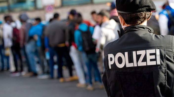 Polizist von hinten mit Schriftzug Polizei auf der Uniformjacke vor eine Schlange mit Flüchtlingen