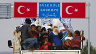 Offener Lastwagen mit Flüchtlingen unter einem türkischen Autobahnschild mit der Aufschrift Güle Güle, Goodbye