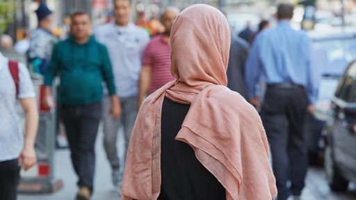 Muslimin mit Kopftuch steht mit dem Rücken zur Kamera auf einem Bürgersteig
