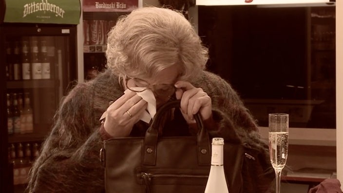 Frau Karger wischt sich mit einem Taschentuch die Tränen ab. Im Vordergrund ein Glas Proseco und die dazugehörige Flasche