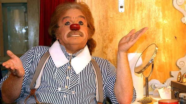 Bernhard Paul als Clown geschminkt. Er trägt ein blauweißes Hemd mit weißem Kragen und eine grau karierte Hose.