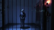 Ein Mann im Anzug in einem blau ausgeleuchteten Raum hinter einem Gitter