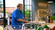 Björn Freitag kocht in einer Küche und hält die Pfanne. 