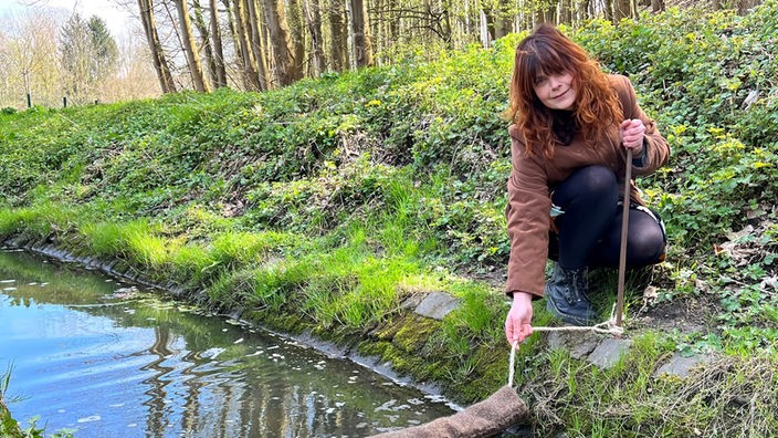 Janine steht im Wald an einem Fluss und montiert etwas im Wasserstrom