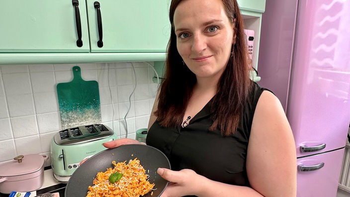 Eine Frau zeigt stolz ihr selbstgemachtes Abendessen in die Kamera, sie steht in der Küche