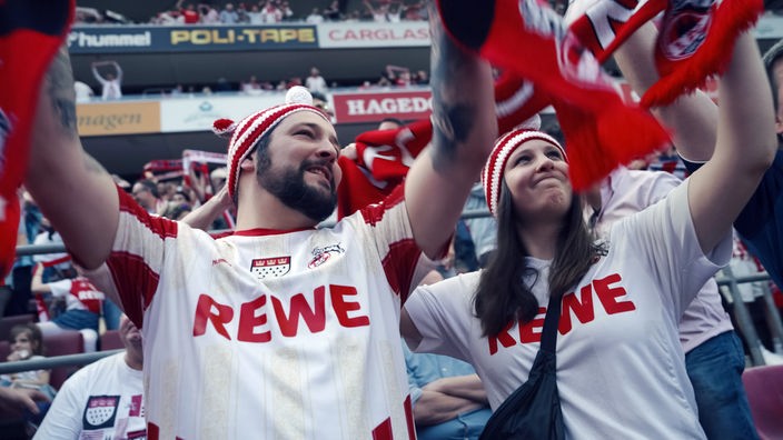 Ein Mann und eine junge Frau in Trikots des FC Köln stehen im Stadion und jubeln