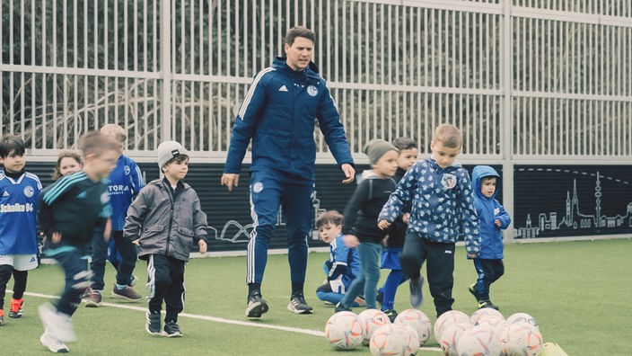 Ein Trainer in Schalke-Trainingsanzug mit vielen Kinder auf dem Fußballplatz