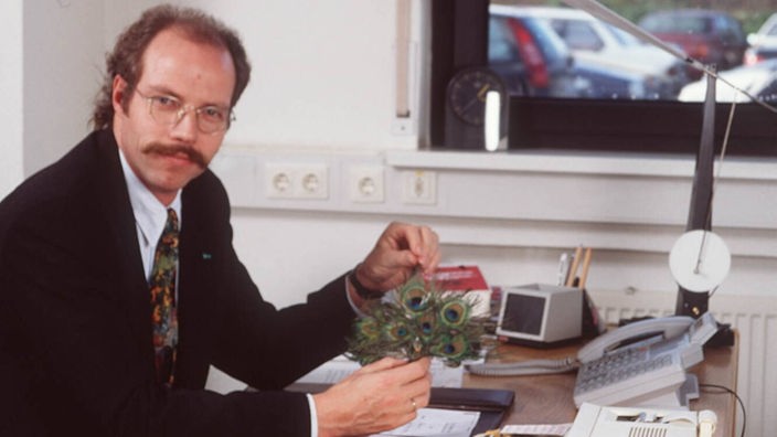 Foto aus den 90er Jahren: Ein Mann sitzt am Schreibtisch, hält einen Spielzeug-Pfau in der Hand