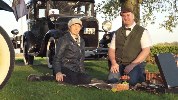 Ein Mann und eine Frau picknicken vor einem Oldtimer.