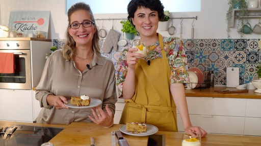 Yvonne Willicks und Theresa Knippschild stehen in einer Küche mit Kuchen und Orangencreme in den Händen.
