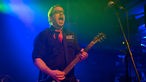 Gitarrist von "The Chuck Norris Experiment" spielt mit weit aufgerissenem Mund im blau-grünen Licht.