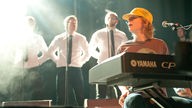 Jonas Nielsen von der Band Kakkmaddafakka spielt Keyboard. Im Hintergrund wartet ein Männerchor auf seinen Einsatz.