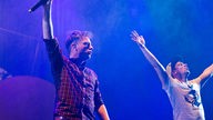 Zwei Sänger reissen in blauem Bühnenlicht die Arme hoch