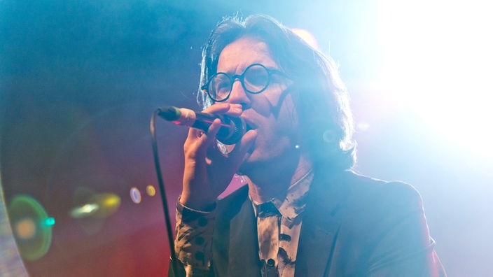 Der Sänger der Band "Spector" trägt eine runde Brille und ein Hemd mit einem 70er/80er Jahre Print unter einem grauen Sacko und hat lange Haare.