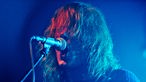 Mann, dem seine langen Haare ins Gesicht fallen, singt in ein Mikrofon im blauen Licht.