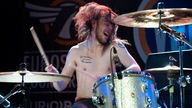 Drummer Pete Flett wirbelt die Drumsticks, seine langen Haare fliegen umher. Er spielt bei nacktem Oberkörper und hat mehrere Tatoos.
