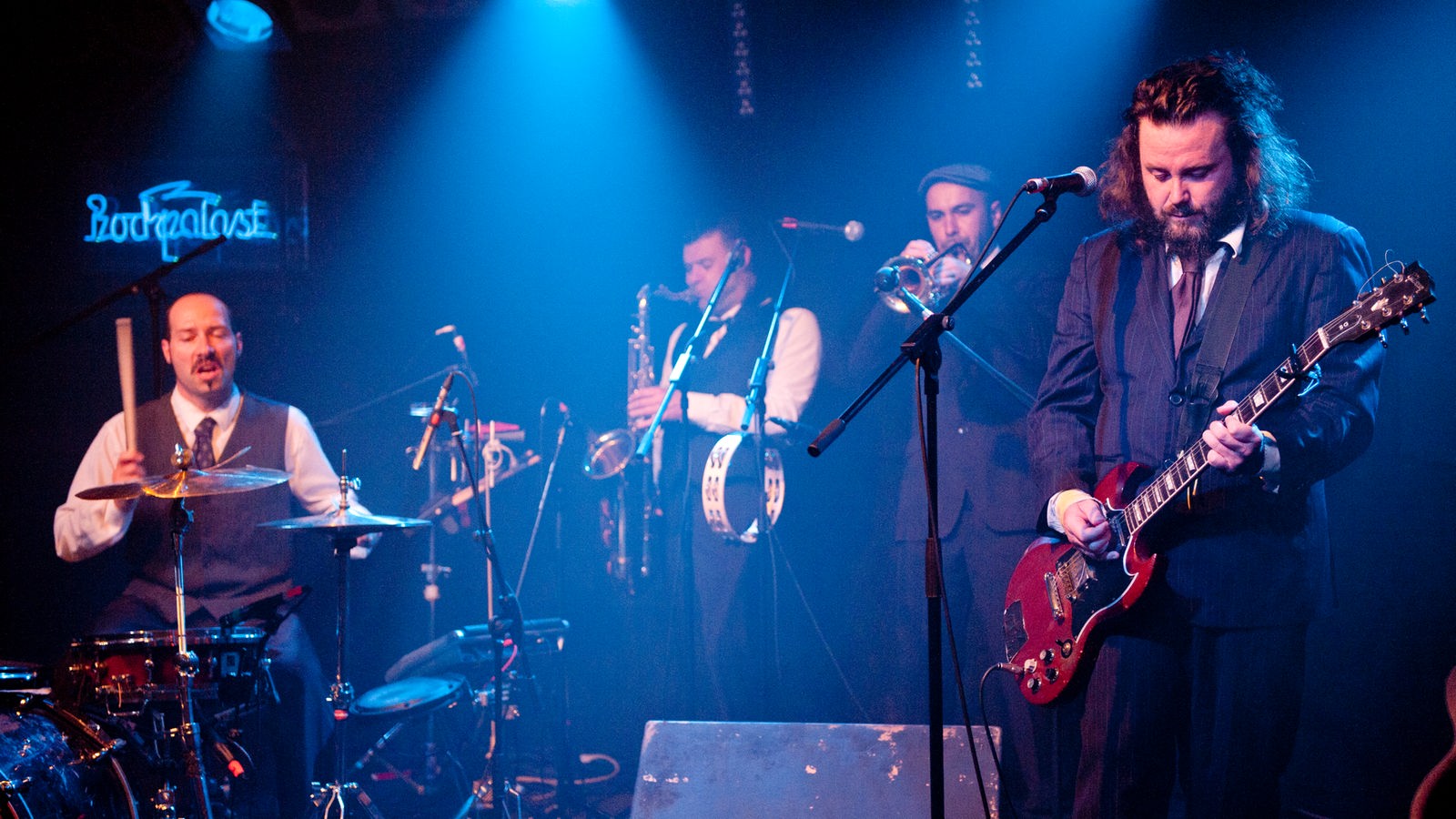 Vier Männer mit Schlagzeug, Saxophon, Trompete und Gitarre im blauen Scheinwerferlicht