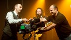 Der Schlagzeuger hält gemeinsam mit einem Kameramann eine Filmkamera auf der Bühne. Im Hintergrund steht der Sänger im gelben Licht.