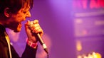 Sänger singt mit geschlossenen Augen in das Mikrofon in seiner Hand. Die Bühne ist violett beleuchtet.