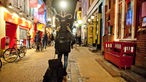 Person mit Gitarrentasche auf dem Rücken und Koffer in der Hand von hinten in einer beleuchteten Gasse in Groningen.