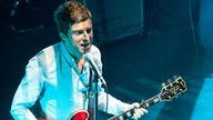 Noel Gallagher steht mit seiner roten Semi-Akustik Gitarre am Mikrofon und wird von oben angeleuchtet.