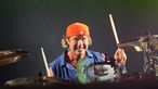 Schlagzeuger mit orangener Schirmmütze an seinem Schlagzeug; grinst in die Kamera