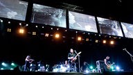 Die Bühne in Makro-Ansicht; darauf die Band, ein Mensch mit Geige, ein Schlagzeug, ein Gitarrist und Sänger und ein Bassist; über der Bühne sind Scheinwerfer und darüber mehrere Bildschirme, die helle Effekte anzeigen