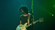 The Zutons: Mann mit Locken steht im grünen Bühnenlicht, hät eine Gitarre und schaut zur Seite