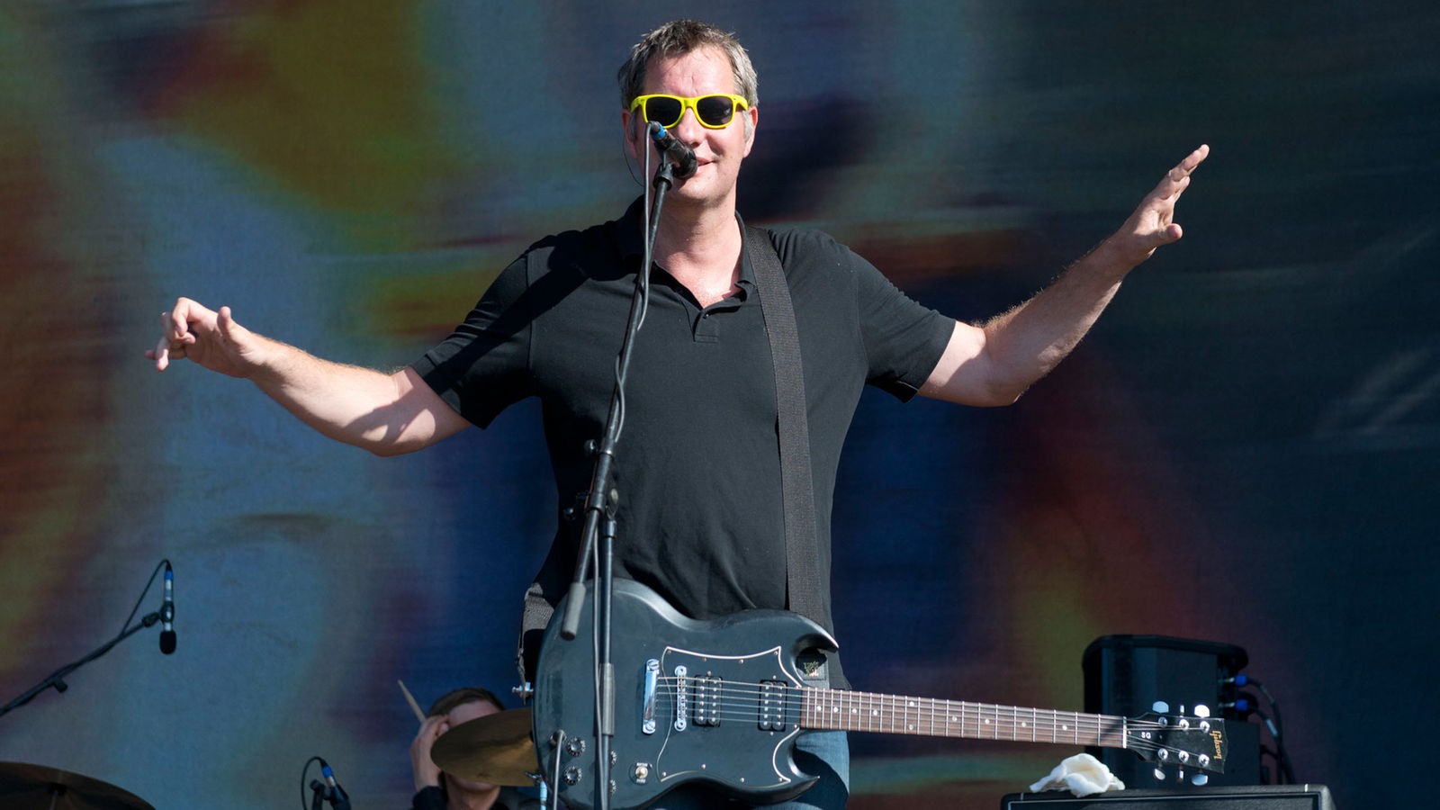Mann mit gelber Sonnenbrille, umgehängter Gitarre und schwarzem T-Shirt breitet seine Arme aus.