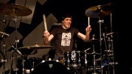 Der Drummer von Skip The Use mit schwarzem T-Shirt und schwarzer Kopfbedeckung zeigt vollen Einsatz an seinem schwarzen Drumkit.