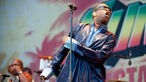 Der Sänger und Komponist Youssou N'Dour trägt eine Sonnenbrille und einen kunstvoll verzierten Regenmantel. Er dreht seinen Kopf nach links.