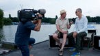Kameramann filmt zwei Männer, die am Seeufer auf Kästen sitzen und miteinander reden
