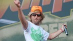 Mann mit blonder Perücke, orangem Hut und Sonnenbrille tanzt mit Mikrofon in der Hand