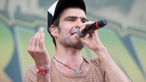 Mann mit Kappe singt mit gehobener Hand in ein Mikrofon