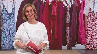 Das Bild zeigt Yvonne Willicks im Servicezeit-Fernsehstudio. Im Hintergrund ist eine Kleiderstange mit magenta-farbenden Klamotten zu sehen.