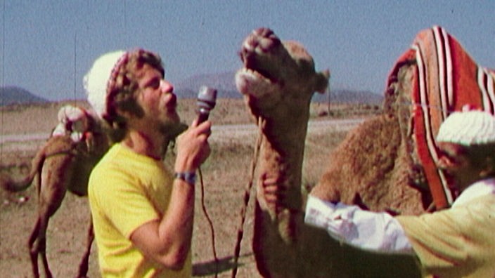 Ein junger Mann hält einem Kamel ein Mikrofon an die Schnauze und tut so, als würde er das Tier interviewen.