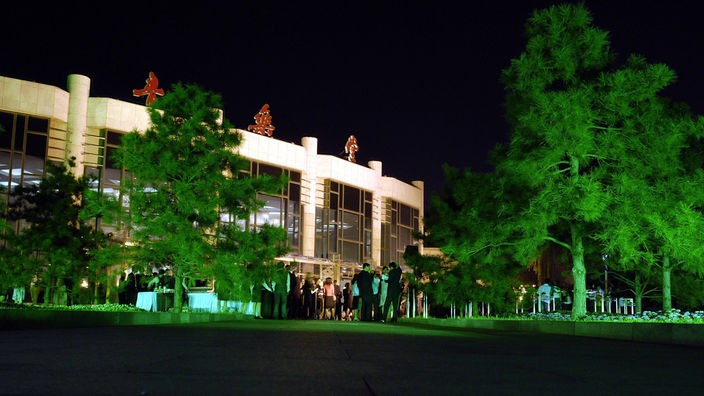 Das Bild zeigt eine Außenaufnahme von einem Konzerthaus in Peking. Davor stehen Bäume, die grün angeleuchtet sind, und mehrere Menschen warten davor auf Einlass. 