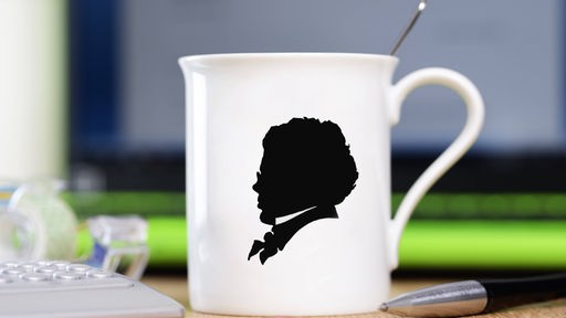 Auf einem Schreibtisch steht ein Kaffeebecher mit der aufgedruckten Silhouette des Komponisten Franz Schubert