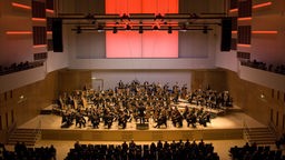 Das Bild zeigt einen großen Konzertsaal. Auf der Bühne sitzen die Duisburger Philharmoniker mit ihren Instrumenten. 