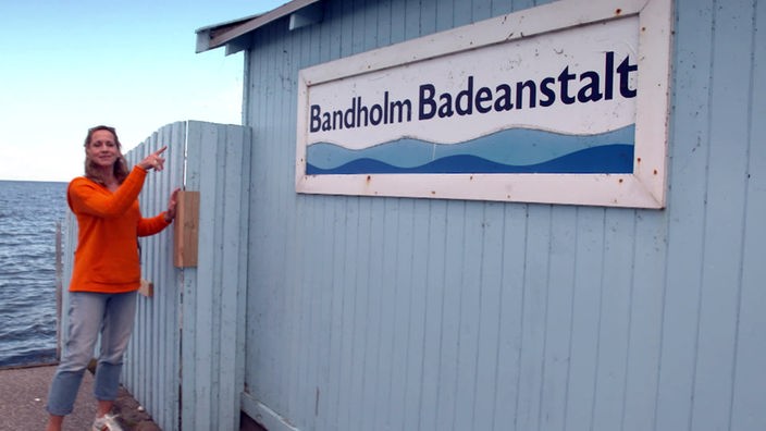 Tamina Kallert steht an einem blauen Bretterzaun mit der Aufschrift "Bandholm Badeanstalt"