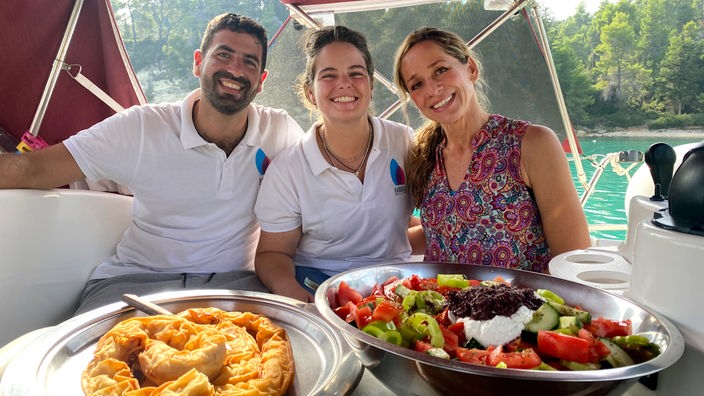 Tamina Kallert (r) mit Antonis Marakas und Alexandra Maraka (m) auf einem Segelboot, vor ihnen Platten mit griechischen Speisen