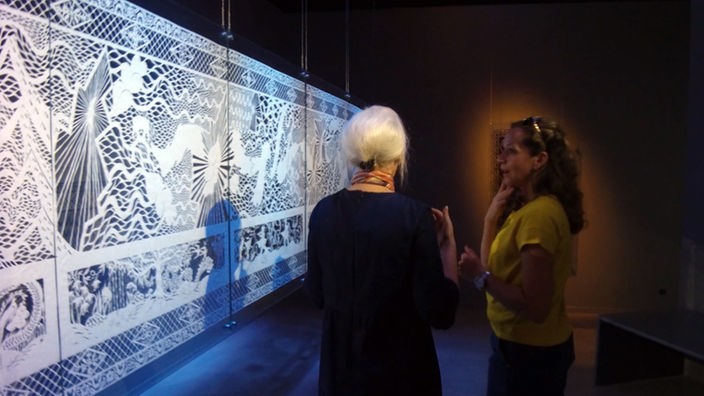 Tamina Kallert (r) und Karen Bit Vejle stehen vor einem großen aus Papier angefertigten Scherenschnitt