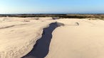 Große Sandfläche mit einem steilen Abbruch, im Hintergrund Küste mit Meer
