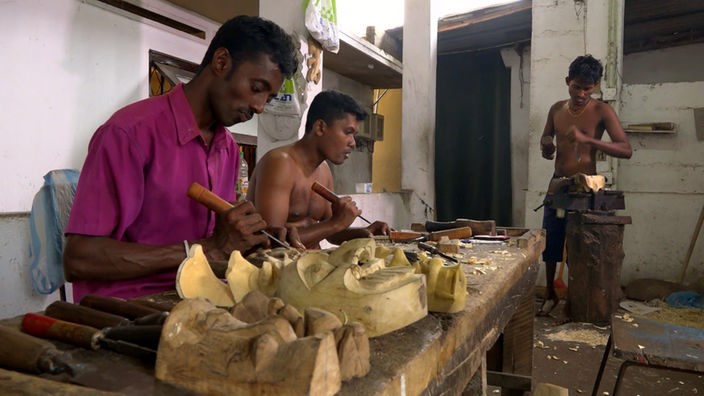 Drei dunkelhäutige Männer arbeiten mit Stechbeiteln an Holzmasken