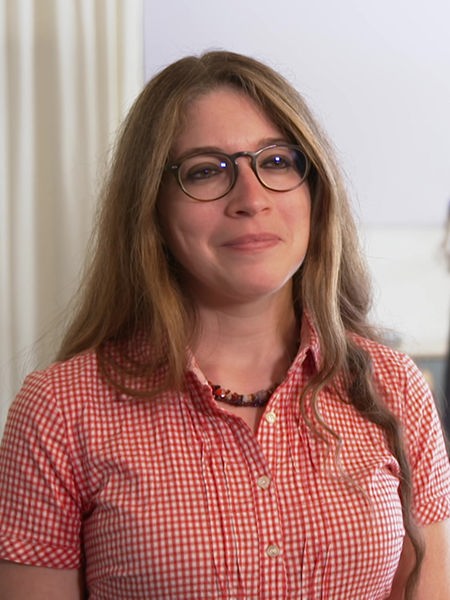 Frau in einem karierten Kurzarmhemd mit langen braunen Haaren und Brille blickt rechts an Kamera vorbei